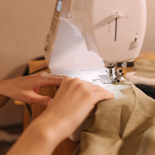 Sichtbar ist eine Nähmaschine und zwei Hände die einen beigen Leinenstoff nähen